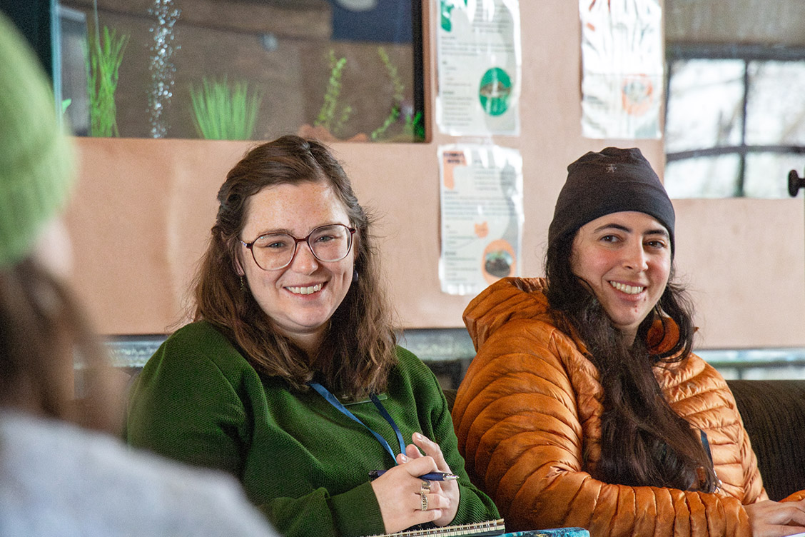 Two people sit indoors and smile at another person.